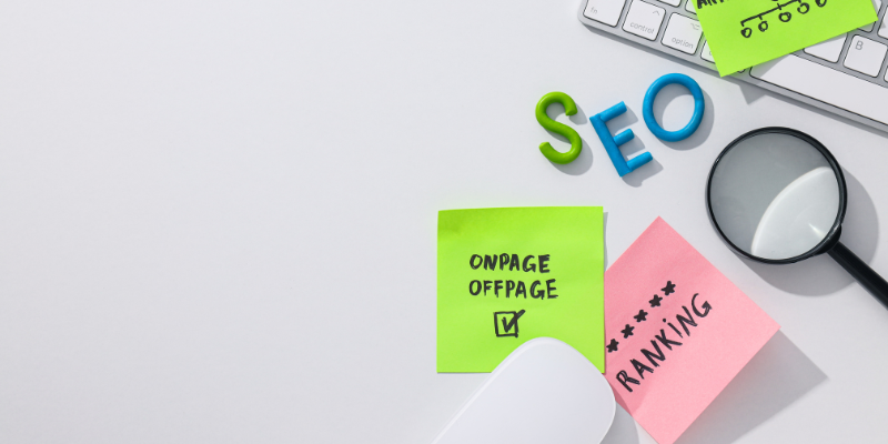 A white table with a keyword and magnifying glass on it. The letters SEO are on the table along with Post-It notes that say on page, off page, and ranking.