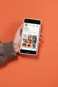 A person holding up a cellphone with a peachy orange background. The phone screen shows an instagram account.
