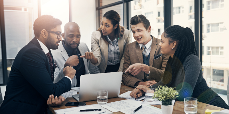 A group of business people at a conference table, talking and pointing to a laptop.