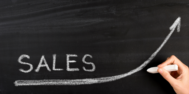 A black chalkboard with someone writing, "sales" and an upward arrow in white chalk on it.