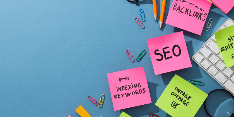 A blue table top with paperclips, keyboard, and pencils. The post it notes say things like, SEO, Backlinks, Indexing Words and Onpage Offpage.