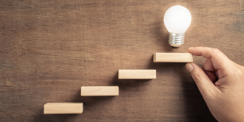 Wooden steps leading up to a lit lightbulb.